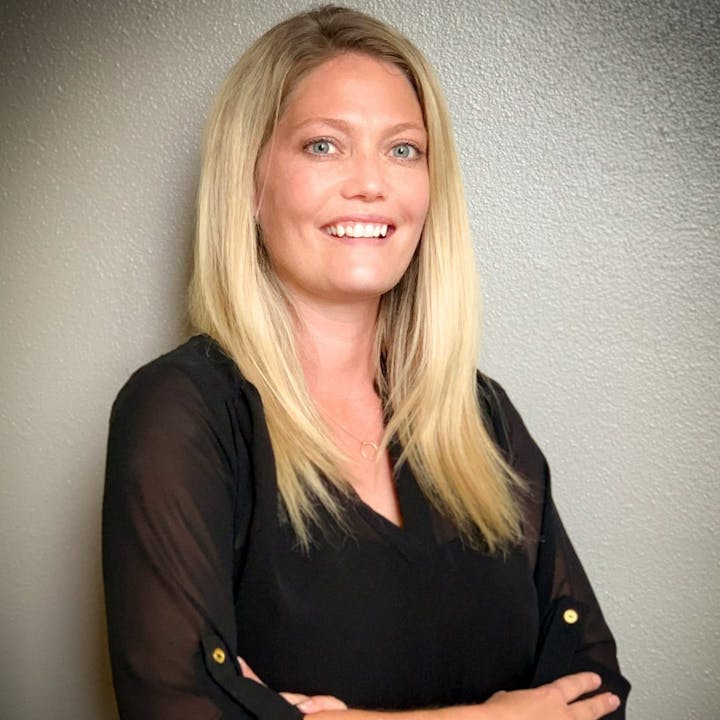 A woman with long blonde hair and a black blouse smiling, standing against a plain wall.