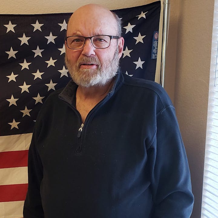 A bearded man in glasses stands in front of a U.S. flag indoors.