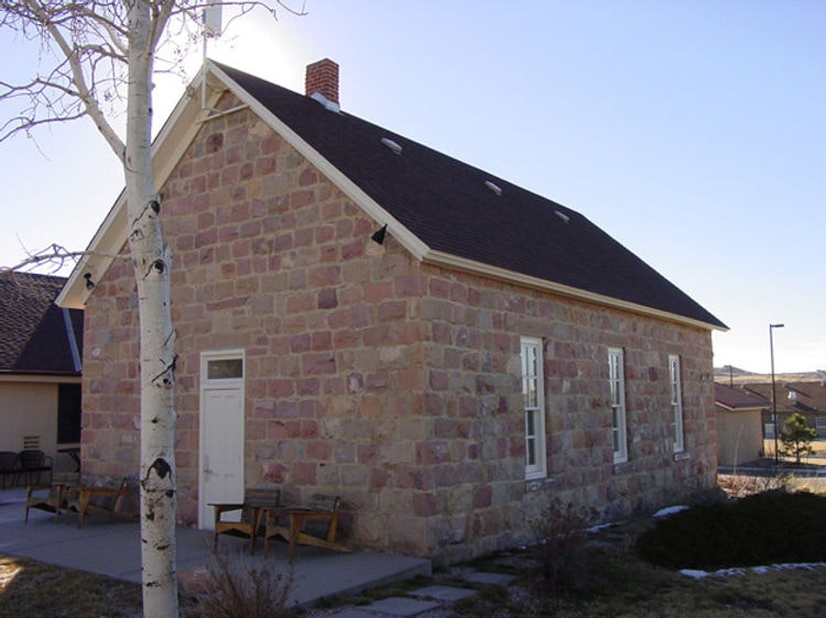 A small stone building with white trim and a sloped roof under a clear sky, with a tree and benches nearby.
