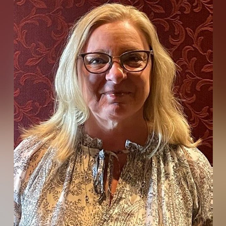 A person with glasses and long blonde hair, wearing a patterned blouse, stands against a dark, textured background.