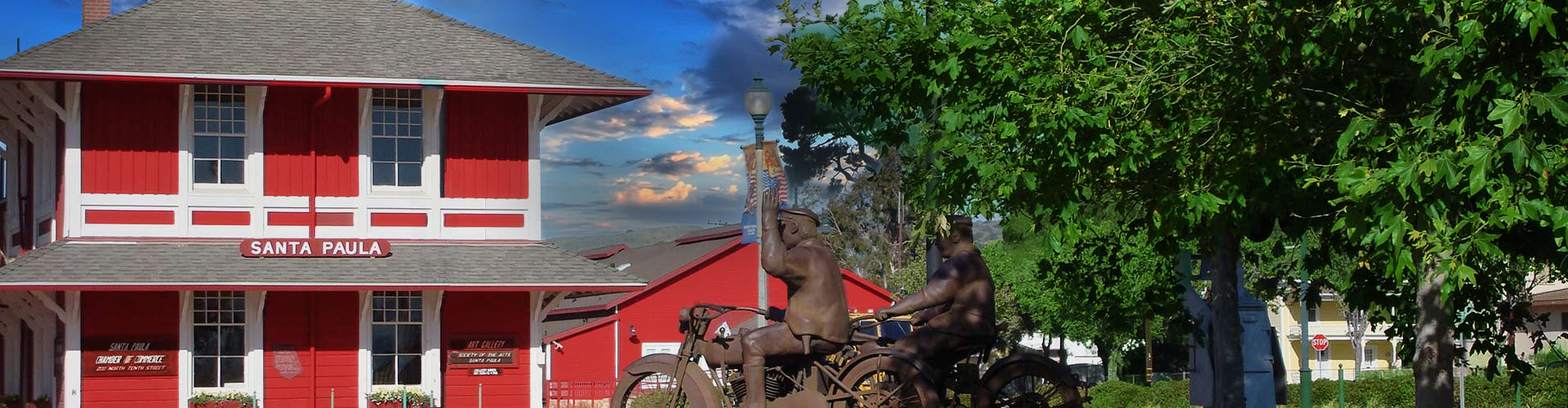 Red and white Santa Paula building, bronze sculpture of cyclists, trees, and a blue sky with clouds.