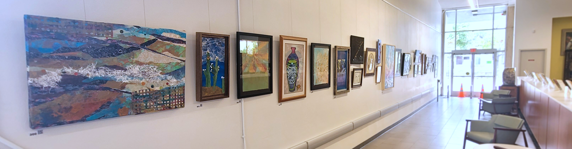 A hallway with a series of paintings hanging on the wall, two chairs, and a doorway at the end.