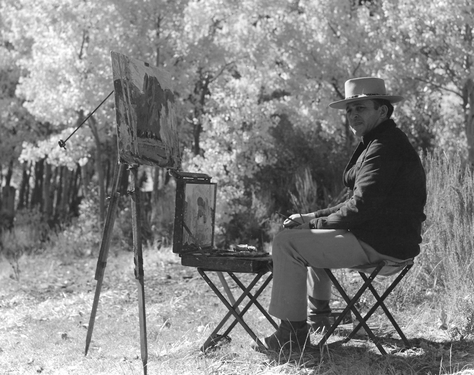 Douglas Shively painting outdoors, sitting beside an easel with a canvas, surrounded by trees.