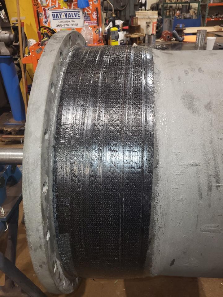 The image shows a close-up of a textured pipe, likely a component in machinery or plumbing, with visible rivets and a workshop background.