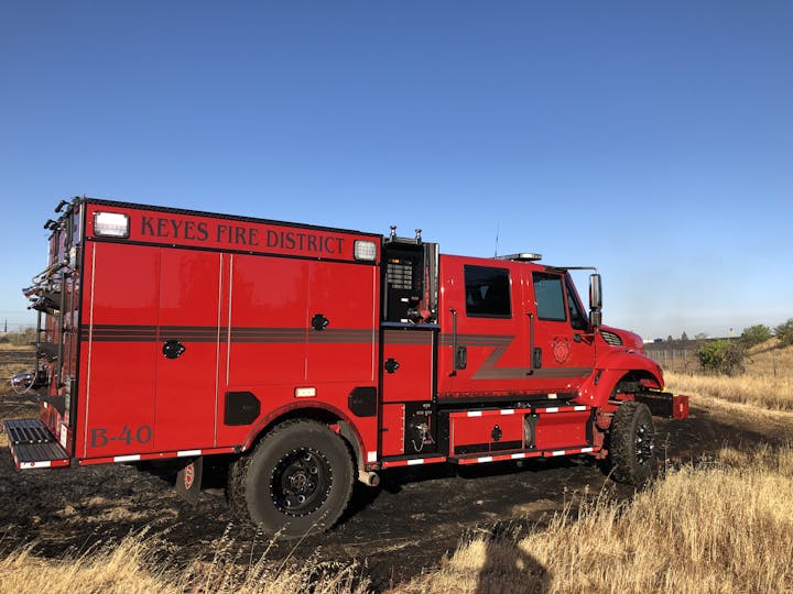 A red fire engine labeled "Keyes Fire District" parked on a dry grass field with a clear blue sky above.
