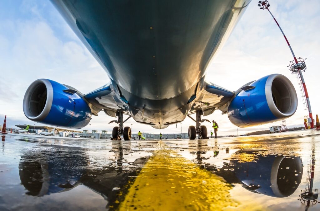 Low-angle view of an airplane's underbelly and engines with ground personnel and reflections on wet tarmac.