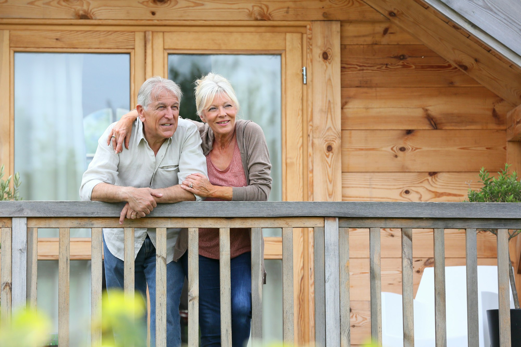 A couple stands on a wooden porch, smiling and leaning on the railing outside a cabin.