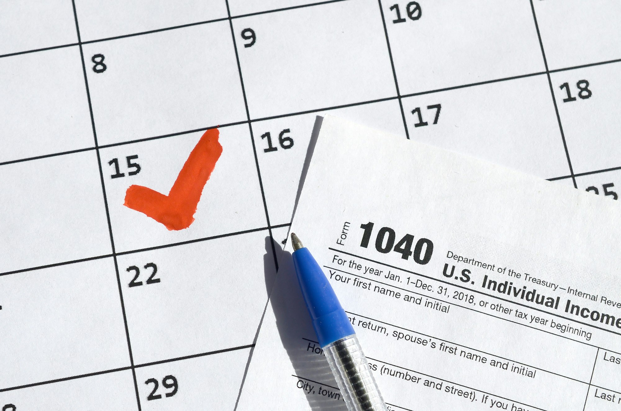 Calendar with a red check mark on the 15th, a 1040 tax form, and a blue pen.