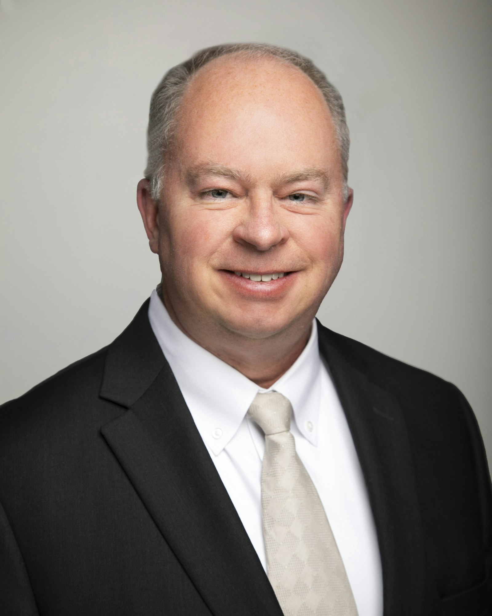 A man in a black suit, white shirt, and light tie, smiling against a plain background.