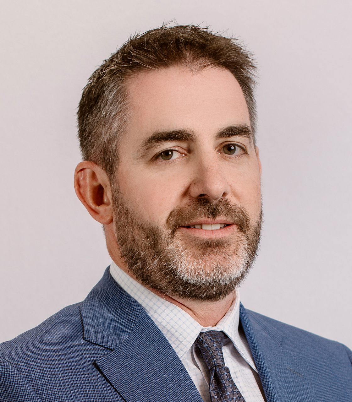 The image features a man with a beard, wearing a light blue suit and tie, against a neutral background.