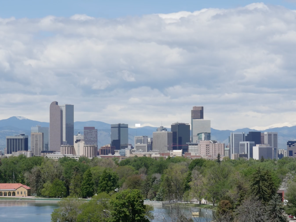 A city skyline with skyscrapers, a lake in the foreground, and mountains in the background under a cloudy sky.