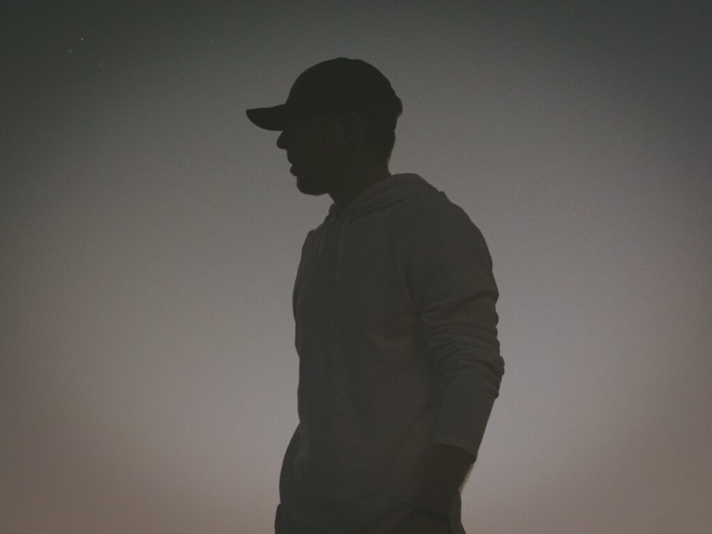 Silhouette of a person wearing a cap and hoodie against a twilight or night sky.