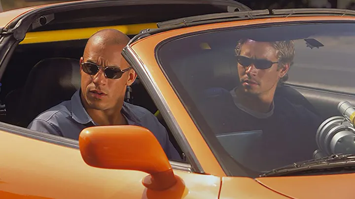 Fast and Furious movie franchise picture, actors in sunglasses sit in an orange sports car, focused and ready for action, surrounded by a sunny environment.