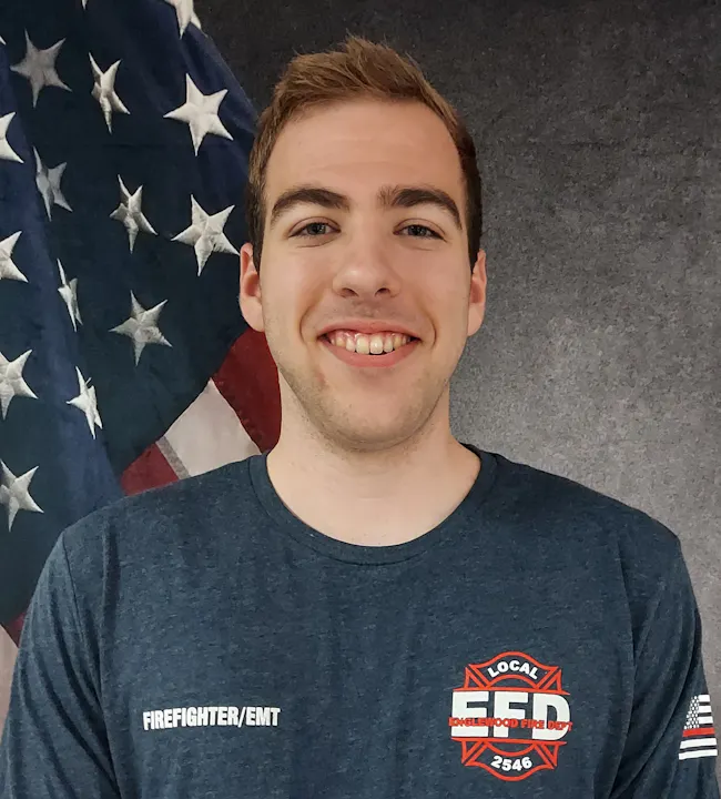 A smiling individual in a firefighter/EMT uniform stands in front of an American flag backdrop.