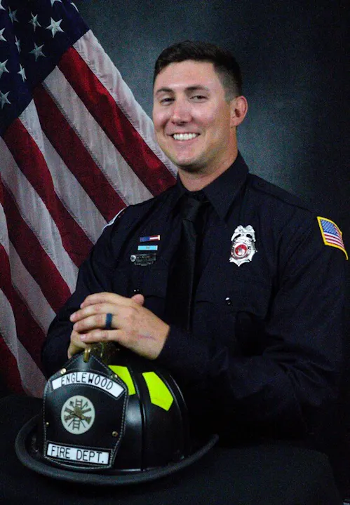 A smiling firefighter in uniform poses with a helmet labeled "Englewood Fire Dept" against a backdrop of the American flag.
