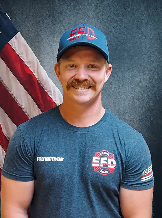 A smiling firefighter/EMT poses in front of an American flag, wearing a cap and shirt with his department's logo.