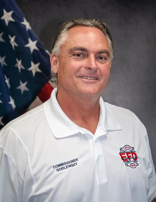 The image features a man in a white polo shirt with "Commissioner Godlewsky" printed on it, smiling in front of an American flag backdrop.