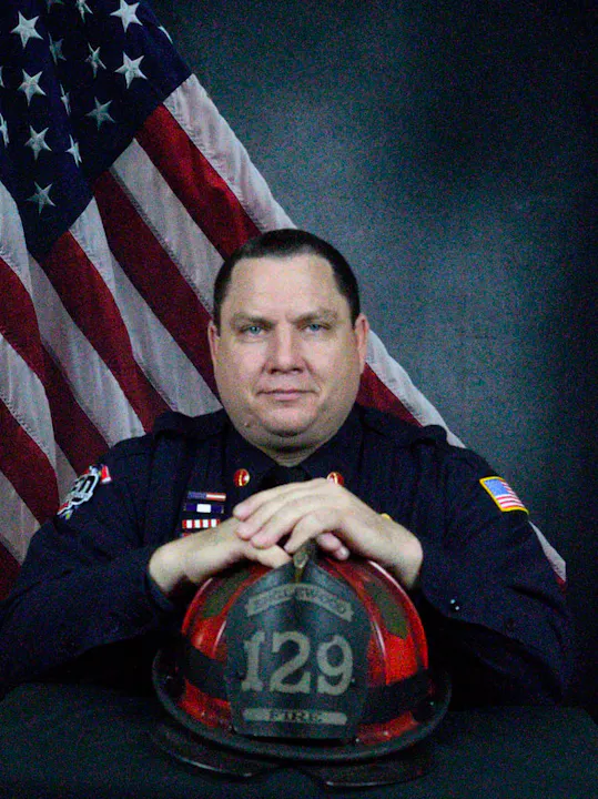 A firefighter in uniform poses with a helmet and an American flag in the background.