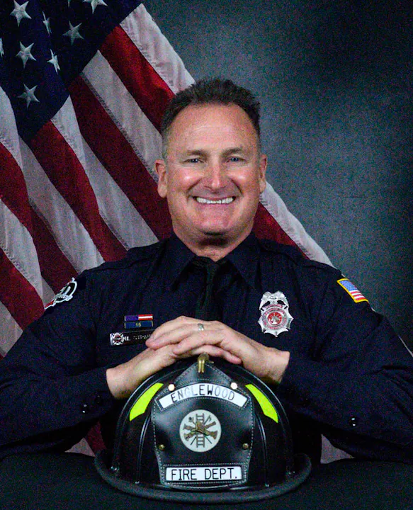 A smiling firefighter is posing in front of an American flag, with a fire helmet labeled "Englewood Fire Dept" on a table.