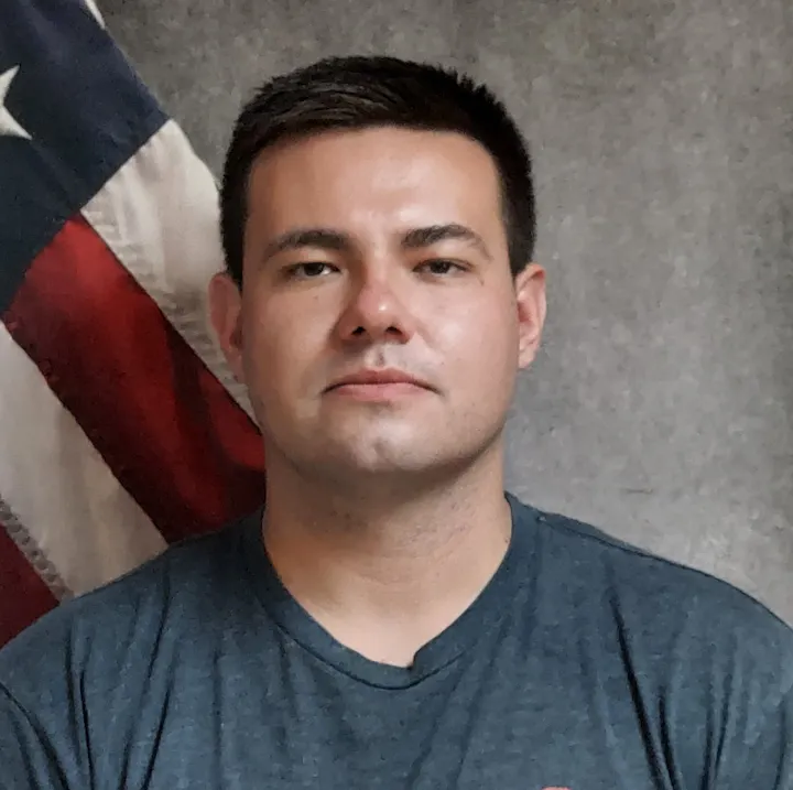 A man in a gray T-shirt with an American flag in the background.