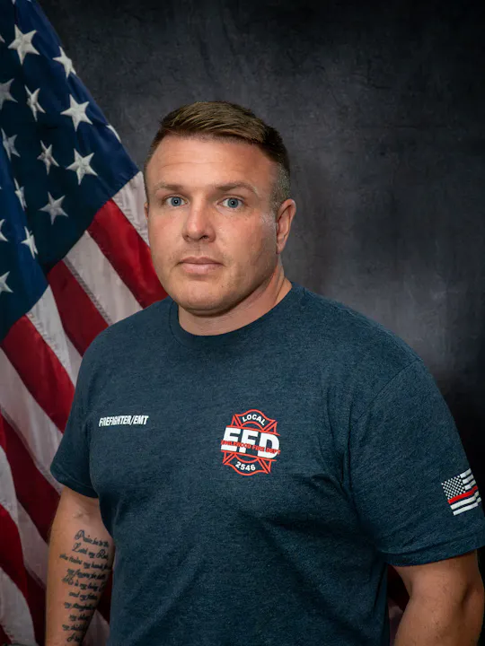 A firefighter/EMT poses for a portrait, with an American flag in the background and a shirt featuring their department’s logo.