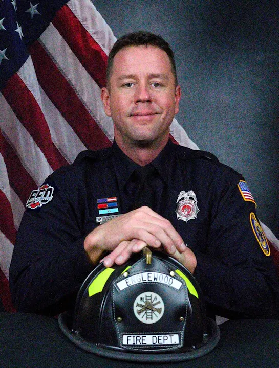 A uniformed firefighter poses with a helmet marked "Englewood Fire Dept" against a backdrop of the American flag.