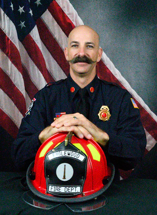 A firefighter in uniform poses with a red helmet labeled "Englewood Fire Dept," against a backdrop of an American flag.