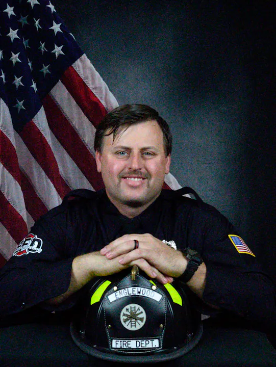 A firefighter poses with his helmet in front of an American flag, smiling confidently.