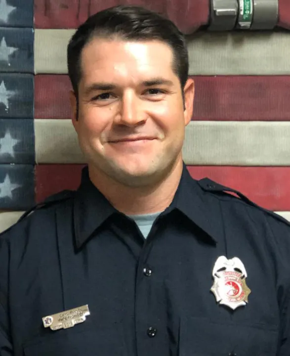 A smiling man in firefighter uniform against an American flag backdrop.