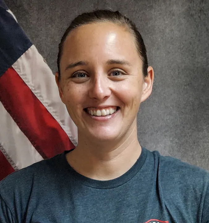 A smiling person in a gray shirt with a portion of the American flag in the background.