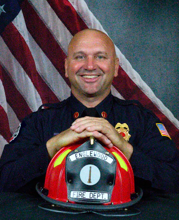 A smiling firefighter in uniform poses with a helmet, against a backdrop featuring an American flag.