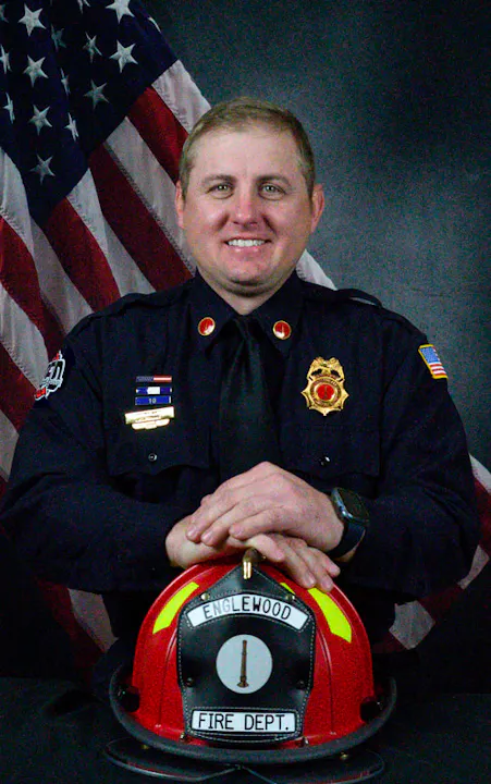 A firefighter poses with a helmet in front of an American flag, showcasing pride and dedication to service.