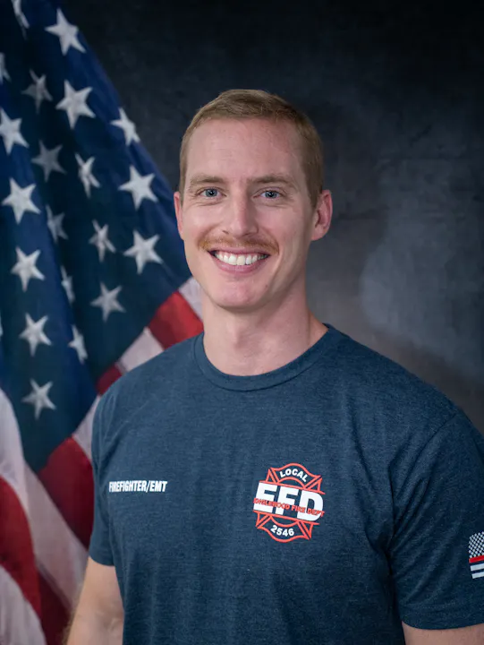 A smiling firefighter/EMT in a uniform shirt stands in front of an American flag backdrop.
