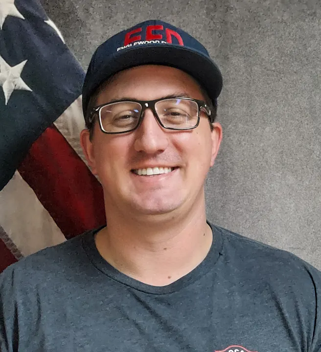 A smiling person wearing glasses and a baseball cap, with a partial view of the American flag in the background.
