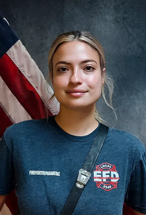 A person in a firefighter/paramedic shirt stands in front of an American flag backdrop, looking directly at the camera.