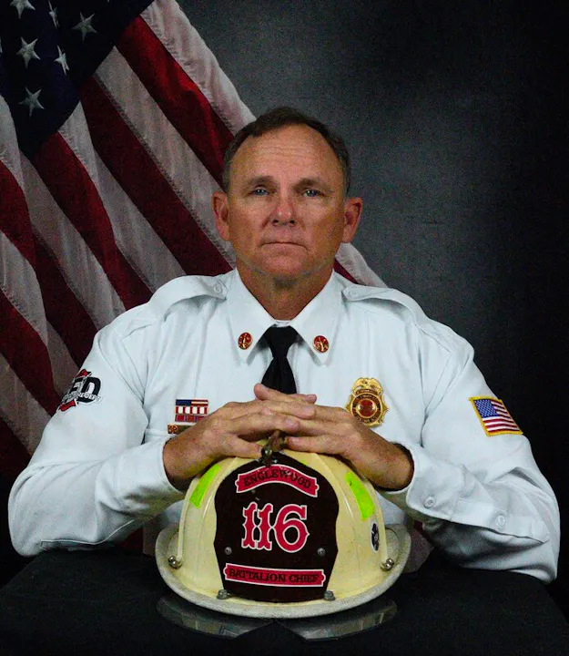A uniformed battalion chief poses with a firefighter helmet in front of an American flag backdrop.