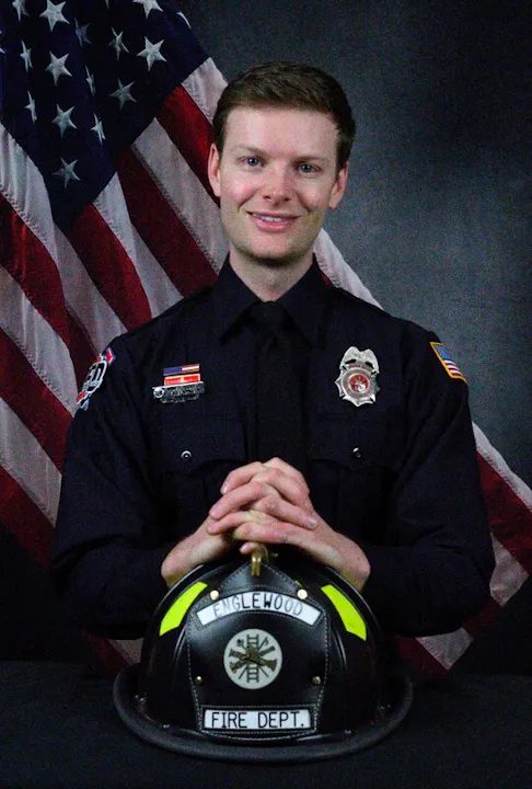 A smiling firefighter in uniform poses with a fire helmet on a table, against a backdrop of the American flag.