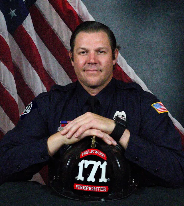 A firefighter poses with a helmet labeled "Englewood 171" against a backdrop of the American flag.