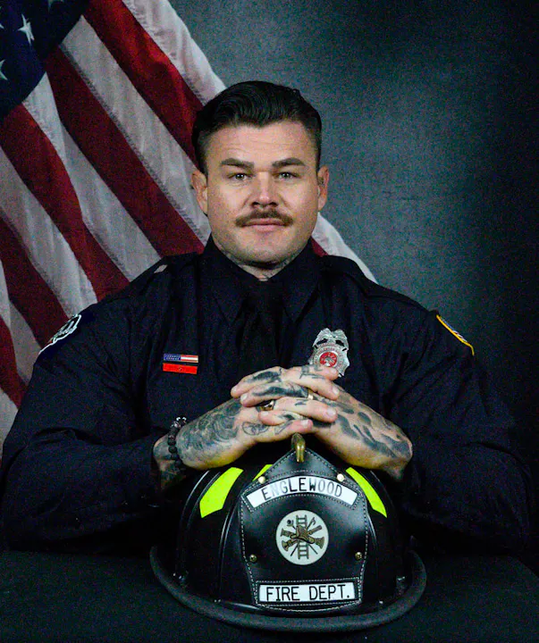 An individual in a fire department uniform with tattoos sits confidently, hands clasped above a helmet, against an American flag backdrop.