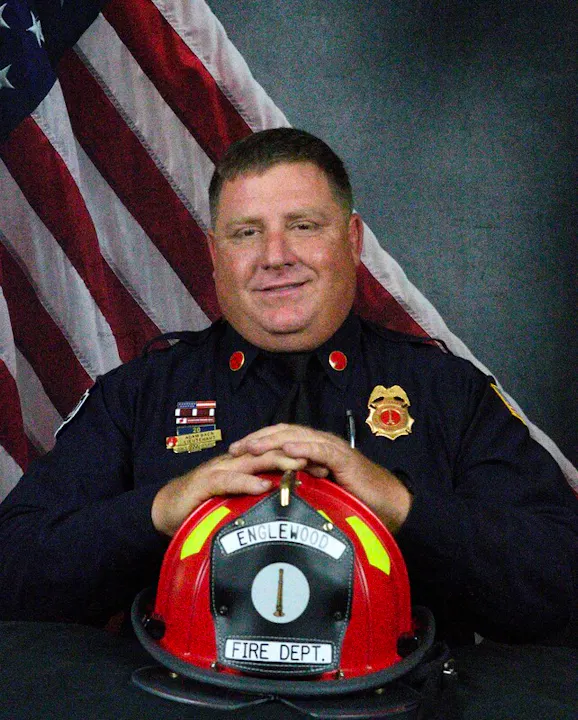 A smiling firefighter poses with a helmet and an American flag backdrop, representing the Englewood Fire Department.