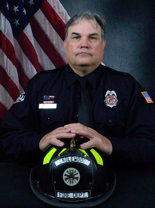 A fire department member poses for a photo with a helmet and an American flag in the background.