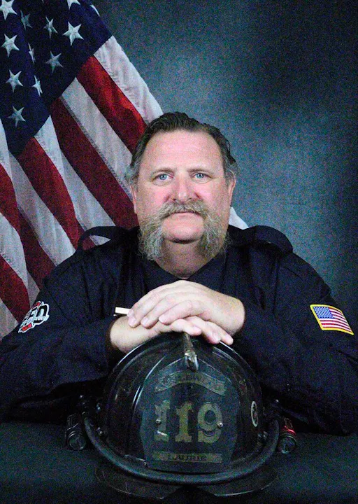A firefighter with a thick mustache poses in front of an American flag, holding a helmet with the number 119.