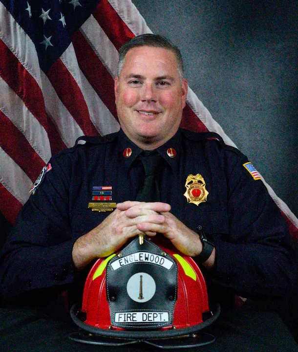 A smiling firefighter in uniform poses with a helmet in front of an American flag background.