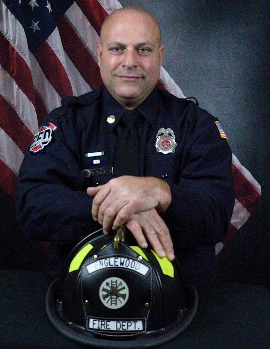 A firefighter in uniform sitting in front of an American flag, holding a helmet.