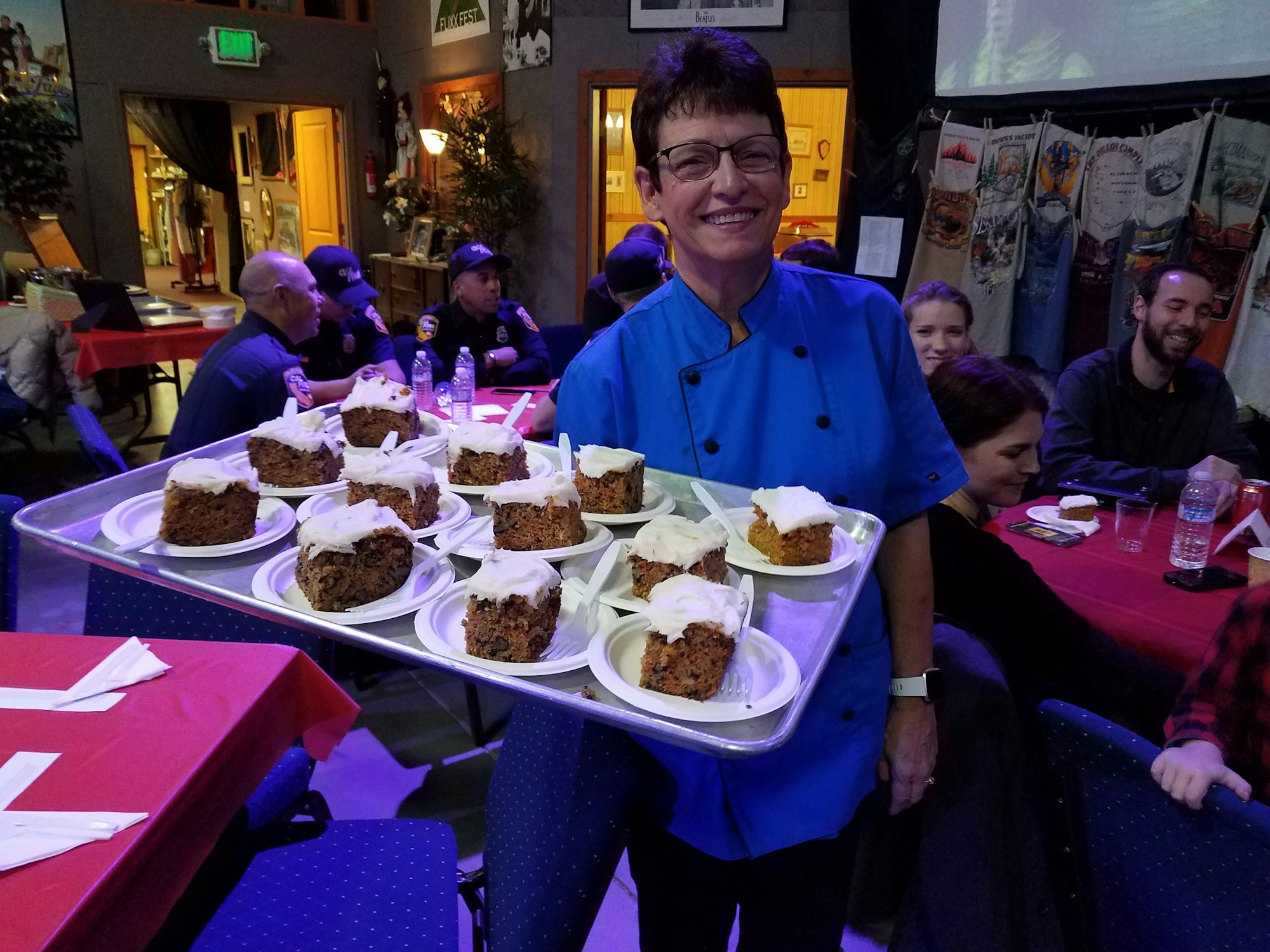 A smiling woman in a blue chef's jacket holds a tray of carrot cake slices at a gathering, with people seated in the background.