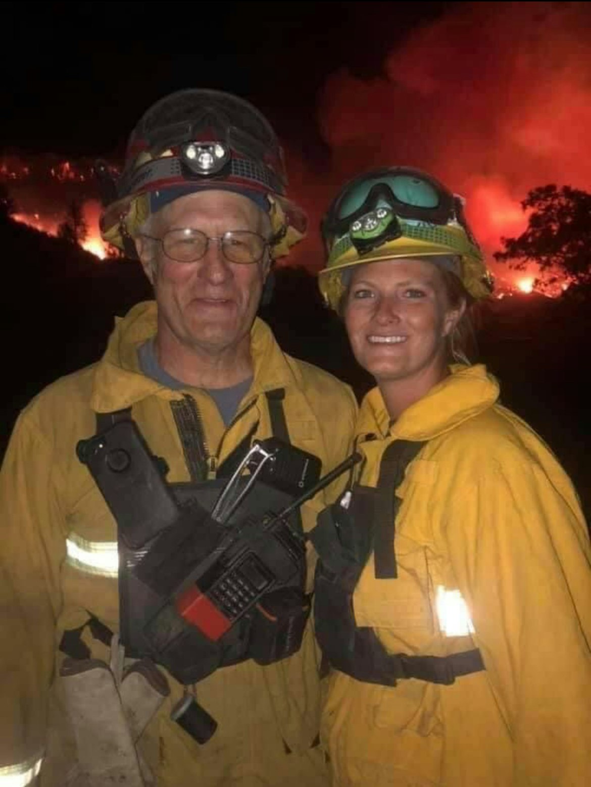 Two firefighters in protective gear with a blazing fire in the background.