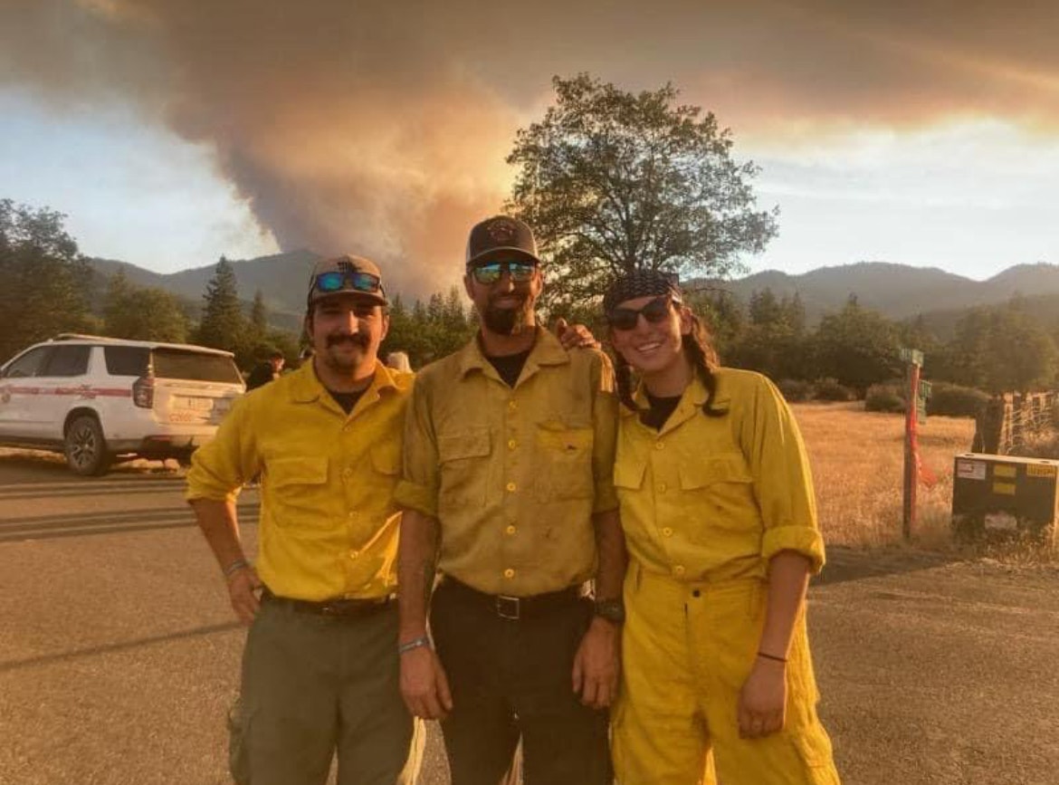 Three firefighters in yellow uniforms with a wildfire smoke plume in the background.