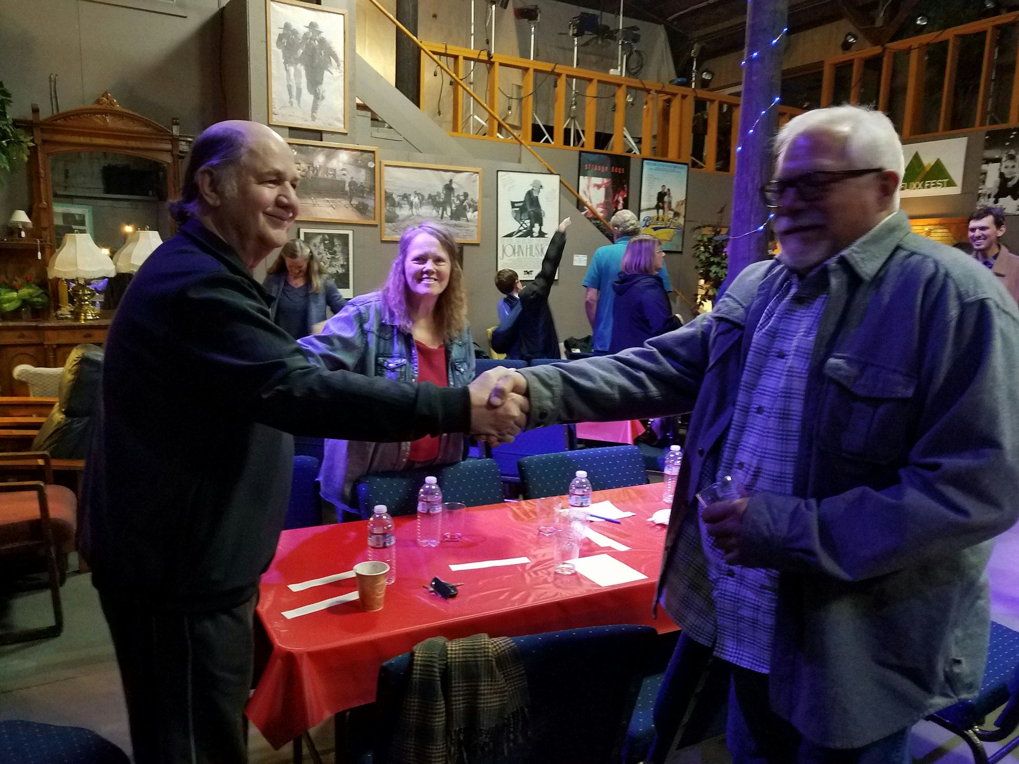 A lively gathering with people shaking hands, seated at a table with water bottles and decorations in a cozy setting.