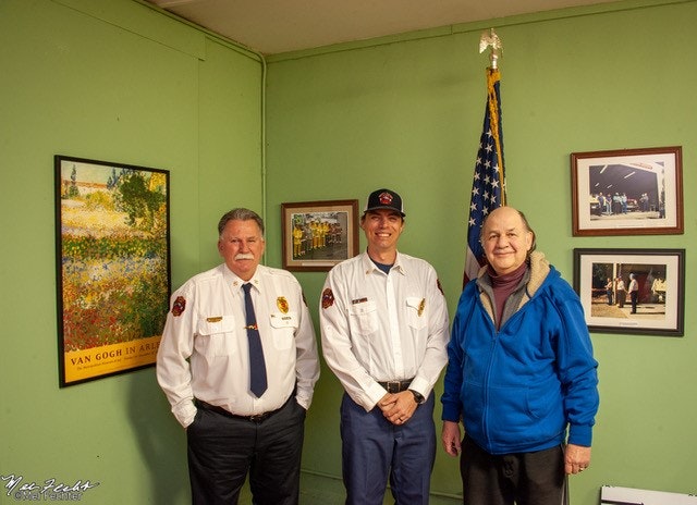 Three individuals, two in fire uniforms and one casually dressed, pose in a room with wall art and an American flag.