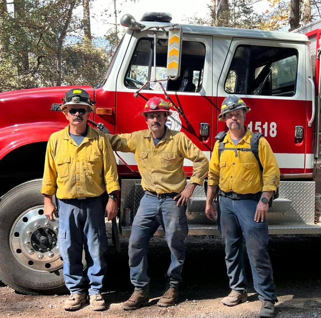 Three firefighters in yellow shirts and helmets in front of a red fire truck, surrounded by trees under a blue sky.